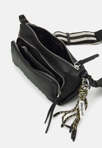 Schwarze Leder-Umhängetasche mit mehreren Reißverschlussfächern, strukturierten Oberflächen und einem gestreiften Tragegurt. Sie verfügt über einen dekorativen Quastenanhänger und Markenlogo.