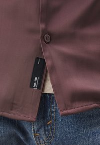 Chemise couleur bordeaux avec une texture lisse, fermeture à un seul bouton et une étiquette en tissu sur l'ourlet, assortie à un jean en denim bleu.