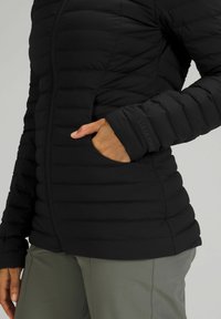 Zwarte pufferjack met een ritssluiting aan de voorkant, ribbeltextuur en zijzakken. Heeft subtiele merkbranding op de manchetten en een getailleerde silhouette.