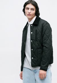 Chaqueta negra acolchada con un cuello de pana, botones a presión plateados y bolsillos laterales, superpuesta sobre una camiseta polo gris y unos jeans azul claro.