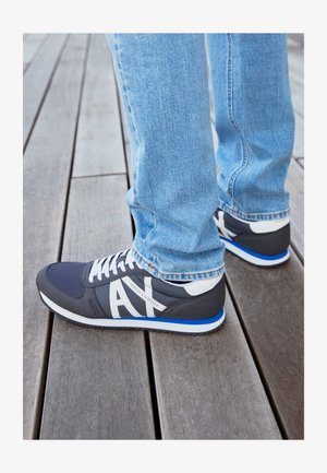 Zapatillas deportivas navy con acentos y logotipo en blanco, con un acabado texturizado. Combinadas con jeans de mezclilla azul claro, sobre un suelo de madera.