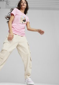 T-shirt di cotone rosa con un design grafico, abbinata a pantaloni cargo beige con molte tasche e sneakers bianche con dettagli viola.