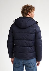 Chaqueta de plumas azul marino con capucha, que presenta un diseño acolchado, puños elásticos y detalles con cordones. El material parece suave y aislante.