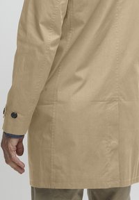 Trench-coat en coton beige avec une coupe droite, des poignets boutonnés et une texture lisse. Doté de poches et de coutures discrètes.