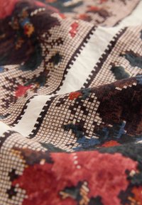 Gros plan d'un tissu avec des motifs géométriques et floraux détaillés en rouge, bleu, marron et blanc, montrant la texture et les plis.