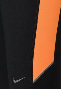 Leggings sportivi neri con una prominente striscia verticale arancione e accenti riflettenti argento, caratterizzati da un piccolo logo Nike vicino all'orlo.