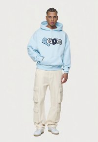 Ljusblå hoodie med svart grafisk text, kombinerad med cremefärgade cargobyxor med flera fickor och vita sneakers.