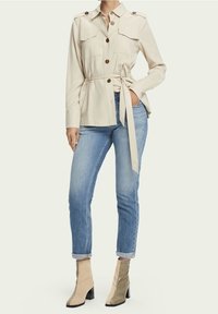 Chaqueta beige de botones con bolsillos frontales, cintura ceñida con cinturón, combinada con vaqueros azul claro arremangados y botas de tobillo beige con estampado de serpiente.