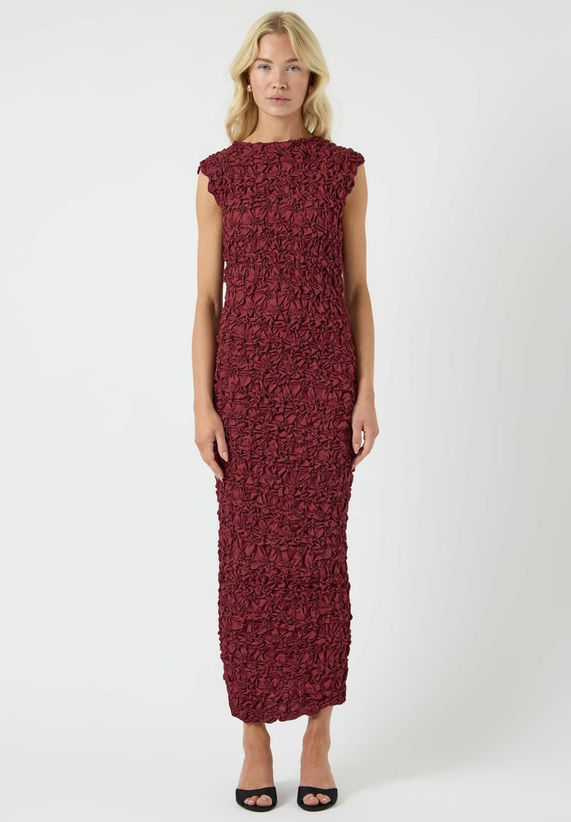 Burgunderfarget ermeløs kjole med strukturert blomstermønster, knelang, tettsittende silhuett og subtil rysjekant rundt halsen, kombinert med svarte høyhælte sko.