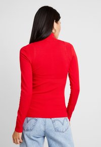 Frau mit langen dunklen Haaren, die goldene Creolenohrringe, einen roten figurbetonten Rippstrickpullover und hellblauen Jeans trägt und sich von hinten zeigt.