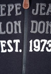 Marinblå huvtröja med dragkedja och dekorativa sömmar. Har texten "PEPE JEANS LONDON" och "EST. 1973" i vitt och grått, med dragkedja av läder.