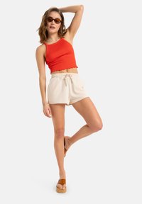 Red ribbed tank top with scalloped hem, beige drawstring shorts, and brown platform sandals. Model poses with one leg raised.