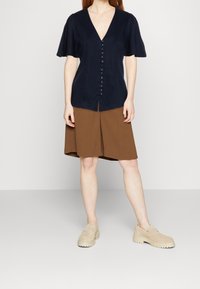 Blusa blu navy a maniche corte con scollo a V e chiusura frontale con bottoni, abbinata a una gonna A-line marrone. Scarpe beige senza lacci con soletta spessa.