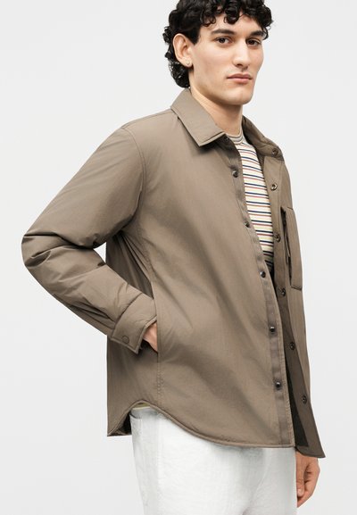 Νεαρό άτομο με σγουρά μαλλιά φοράει ένα μπεζ σακάκι με κουμπιά πάνω από ριγέ πουκάμισο και λευκό παντελόνι, στέκεται με το ένα χέρι στην τσέπη του.