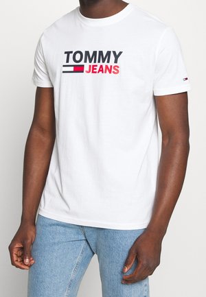 Mies, joka käyttää valkoista lyhythihaista T-paitaa, jossa on "TOMMY JEANS"-logo, ja sinisiä farkkuja, seisoo yksivärisen valkoisen taustan edessä.