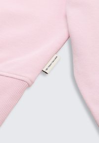 Jasnoróżowa dzianina bluzy z ściągaczami i stębnowanym dołem. Posiada metkę z napisem 'TOM TAILOR' w kontrastowym kolorze.