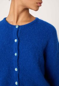 Moteris dėvi ryškiai mėlyną pūkuotą kardiganą su apvaliais šviesiai mėlynais mygtukais, matoma nuo kaklo iki vidurio liemens, žvelgianti iš šono.