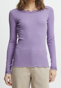 Mujer vestida con una camiseta ajustada de manga larga color lavanda con encaje en el escote y en los puños, combinada con pantalones beige frente a un fondo liso.