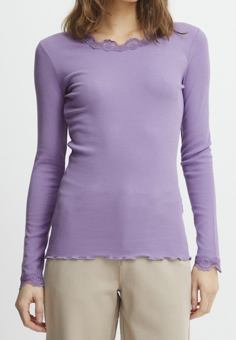 Mujer vestida con una camiseta ajustada de manga larga color lavanda con encaje en el escote y en los puños, combinada con pantalones beige frente a un fondo liso.