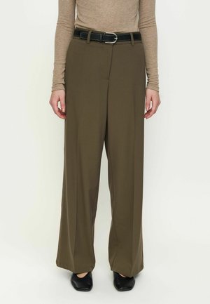 Personne portant un haut beige ajusté à manches longues, un pantalon de costume olive à jambes larges avec une ceinture noire et des chaussures noires, debout devant un fond uni.