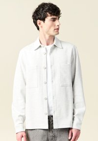 Jasnoszara koszula z długim rękawem i kołnierzykiem, zapinana na guziki, z dwiema przednimi kieszeniami, uszyta z miękkiego materiału, noszona z białym podkoszulkiem pod spodem.