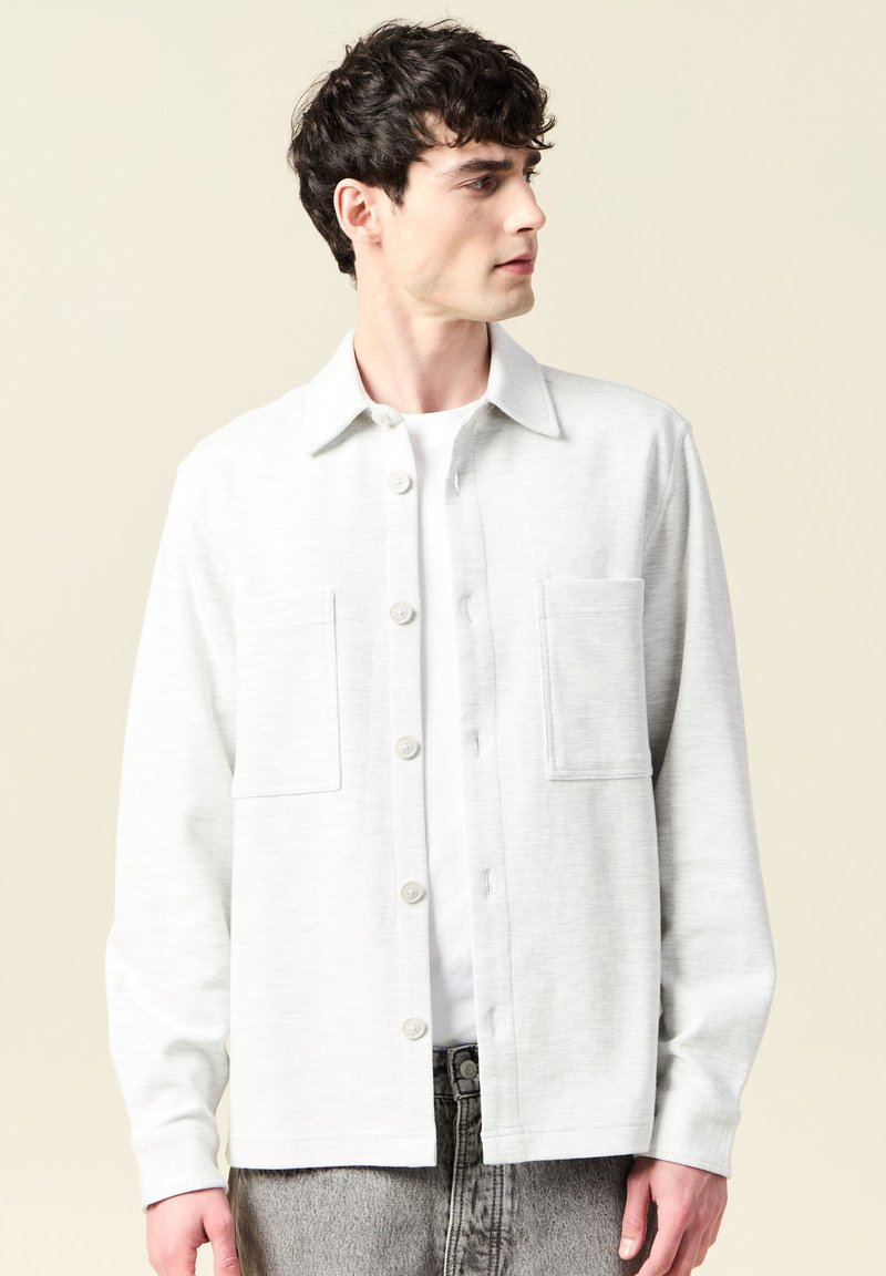 Jasnoszara koszula z długim rękawem i kołnierzykiem, zapinana na guziki, z dwiema przednimi kieszeniami, uszyta z miękkiego materiału, noszona z białym podkoszulkiem pod spodem.