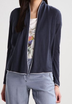 Femme portant un cardigan bleu foncé ouvert sur un t-shirt blanc avec un motif graphique et un pantalon bleu clair.