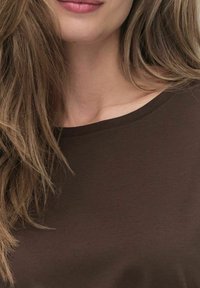 Tissu doux de couleur marron foncé avec un col rond et des manches courtes. Détail de couture le long du col. Cheveux partiellement visibles.