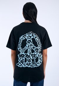 Camiseta de algodón negra con un diseño de signo de paz azul, acentuado con flores y texto, ubicado de manera destacada en la parte posterior.