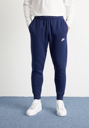 Person iført marineblå Nike joggingbukser og hvide sneakers, stående på blåt tæppe med hænderne i lommerne.