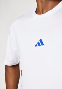 Weißes Sportshirt aus strukturiertem Stoff, mit einem blauen Adidas-Logo und roten Akzenten an den Seiten. Kurze Ärmel und Rundhalsausschnitt.