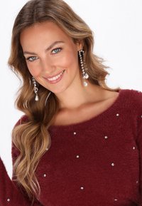 Burgunderfarbener, flauschiger Pullover mit weitem Ausschnitt, der abwechselnd kleine silberne Nieten und Kristallakzente aufweist. Lange Haare teilweise sichtbar.