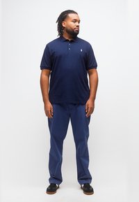 Άνδρας με ντραντλοκς, φοράει μπλε ναυτικό πόλο, παντελόνια στο ίδιο χρώμα και μαύρα αθλητικά παπούτσια, στέκεται και κοιτάζει προς τα πλάγια με φόντο λευκό.