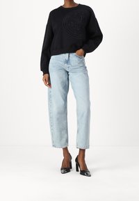 Sudadera negra con logo texturizado, combinada con jeans de mezclilla de pierna recta de talle alto en azul claro. Unas elegantes zapatos de tacón negros brillantes completan el conjunto.