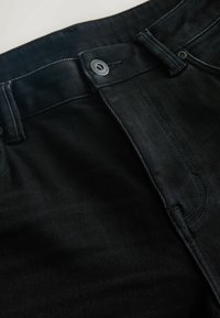 Melnās denim bikses ir ar gludu tekstūru un klasisku piecu kabatu dizainu, metāla pogu aizdari un smalkiem šuvuma elementiem.