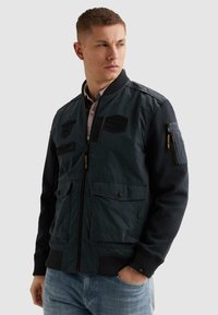 Ung mand iført en mørkeblå arbejdsjakke med patches, lynlås- og knaplommer, står med den ene hånd i jeanslommen og kigger sidelæns.