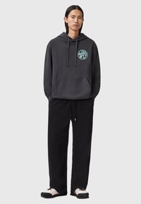 Felpa grigia con stampa grafica circolare sul petto, abbinata a pantaloni da tuta neri e sneakers beige e nere. Vestibilità e stile casual.