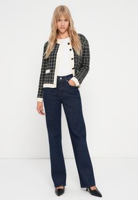 Μαύρο και λευκό καρό πουλόβερ με στρογγυλή λαιμόκοψη και κουμπιά, συνδυασμένο με τζιν ψηλόμεσα σε σκούρο μπλε χρώμα και μαύρες γόβες.