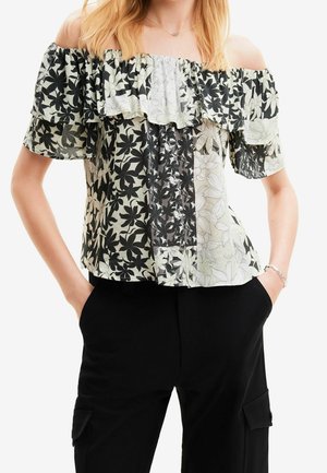 Woman wearing off-shoulder black and white floral ruffled top with short sleeves and black pants with side pockets, hands in pockets.