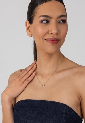 Frau mit dunklen Haaren in einem trägerlosen Denim-Top, die eine goldene Kette mit kleinem Blumenanhänger trägt und sanft ihr Schlüsselbein berührt.