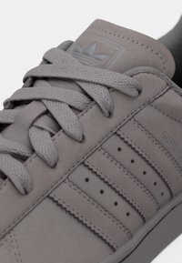 Γκρι σουεντ αθλητικό παπούτσι με επίπεδα κορδόνια, τρεις διάτρητες ρίγες στο πλευρό και ετικέτα με λογότυπο στη γλώσσα. Ομαλή υφή, μινιμαλιστικός σχεδιασμός.