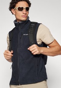 Navy fleece vest with a high collar, zip closure, and two front pockets. Worn over a light shirt with a backpack and sunglasses.