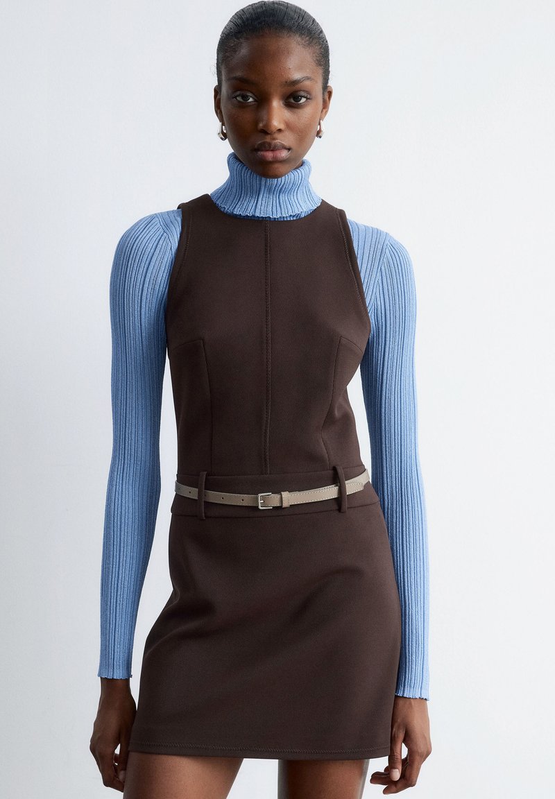 Braunes ärmelloses Kleid mit Seitentaschen, kombiniert mit einem hellblauen gerippten Rollkragenpullover. Ein kontrastierender beiger Gürtel betont die Taille.