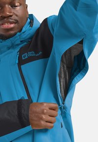 Veste d'extérieur légère bleue avec des accents noirs, des ouvertures de ventilation zippées et une doublure en mesh, avec un logo sur la poitrine.