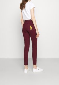 Jeans skinny bourgogne avec un logo polo brodé jaune sur la poche arrière, associés à un t-shirt blanc et des baskets blanches.