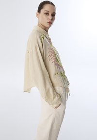 Camisa beige bordada con diseños florales en verde y rosa, con un corte relajado, manga larga y cuello con botones. Combinada con pantalones crema.