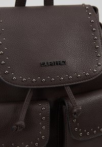 Sac à dos en cuir brun texturé avec détails en métal clouté, fermeture par cordon et poches avant. Logo "SURIFREY" affiché en évidence.