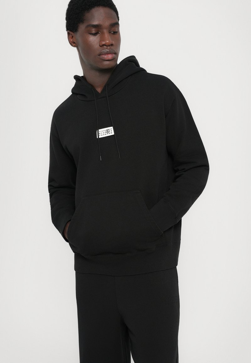 Hombre con sudadera negra con un pequeño logo rectangular blanco en el pecho, manos en el bolsillo delantero, mirando hacia abajo a su izquierda contra un fondo liso.