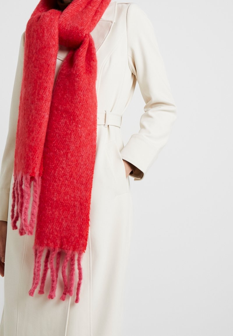 Personne portant un manteau blanc ceinturé avec une épaisse écharpe rouge texturée enroulée autour du cou, tombant avec des franges aux extrémités.