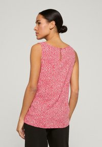 Červený top bez rukávů s bílým květinovým vzorem, detailem klíčové dírky na zádech a uvolněným střihem, vyrobený z lehkého materiálu. Pod ním jsou nošeny černé kalhoty.